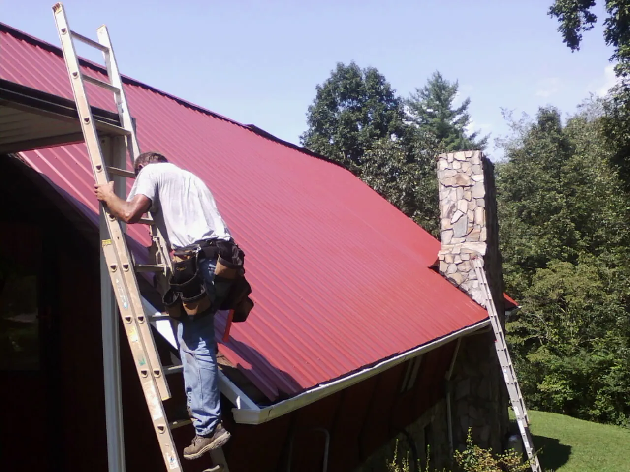 Roofer on a ladder beside a red metal roof and stone chimney — Soffit & Fascia Repair by Roofing Brothers in Bomont